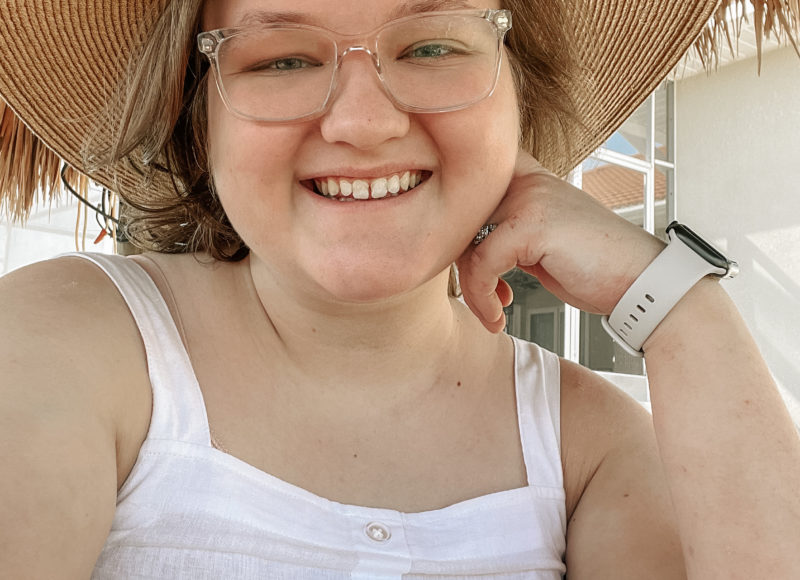 Elliot, a nonbinary person, takes a selfie in a white tank top and tan sun hat